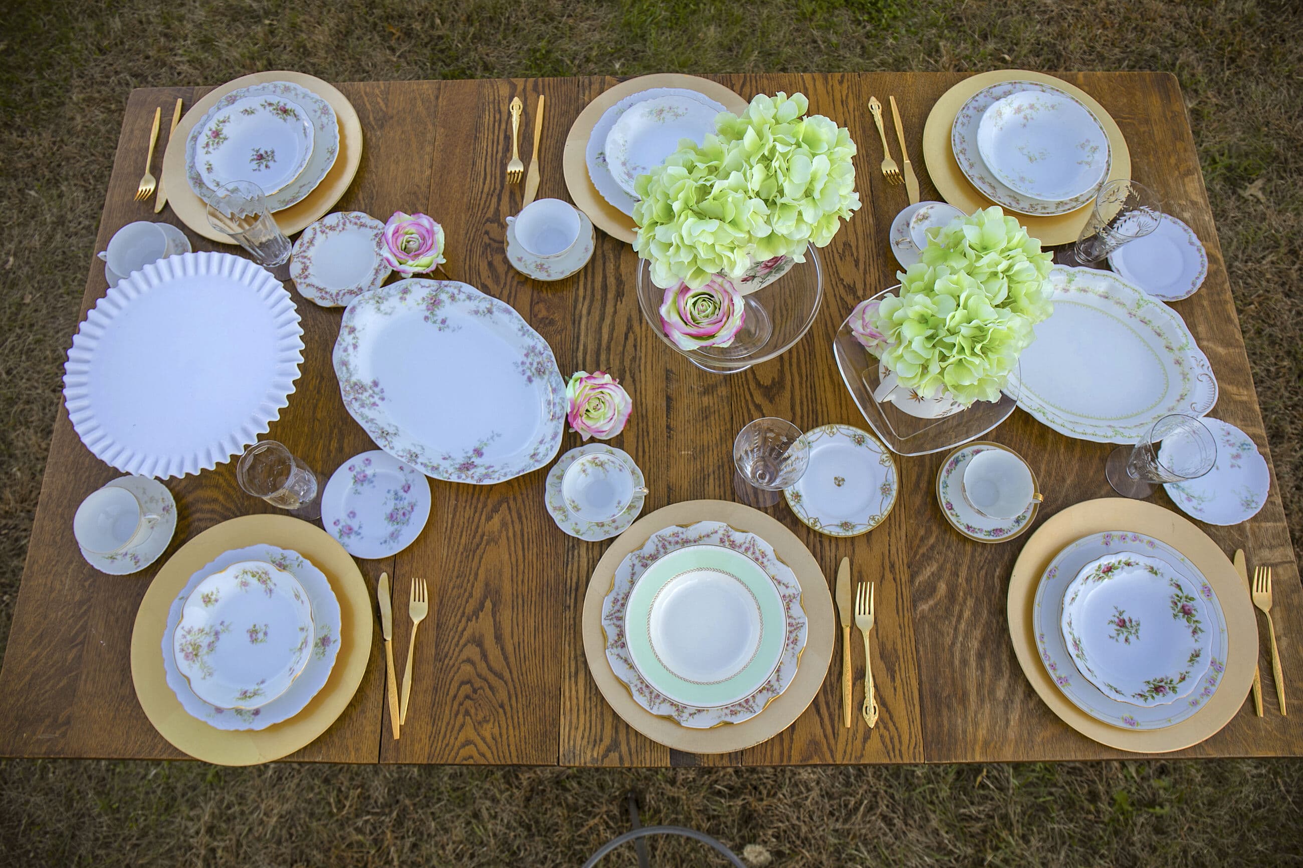 An overhead view of a fully set vintage table: mismatched floral china, milk glass, and green hydrangeas on gold chargers